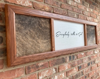Op de afbeelding: Een lange, rechthoekige houten lijst met drie panelen. Het middelste paneel is wit met de tekst "Everybody talks to God" in zwarte cursieve letters. De linker- en rechterpanelen zijn bedekt met bruin en wit koehuid.
