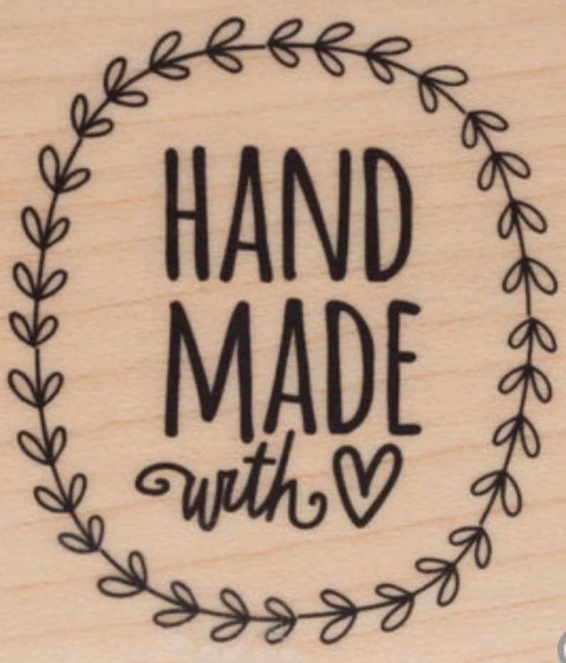 May include: A black and white stamp with a wreath of leaves around the text "HAND MADE with &hearts;".