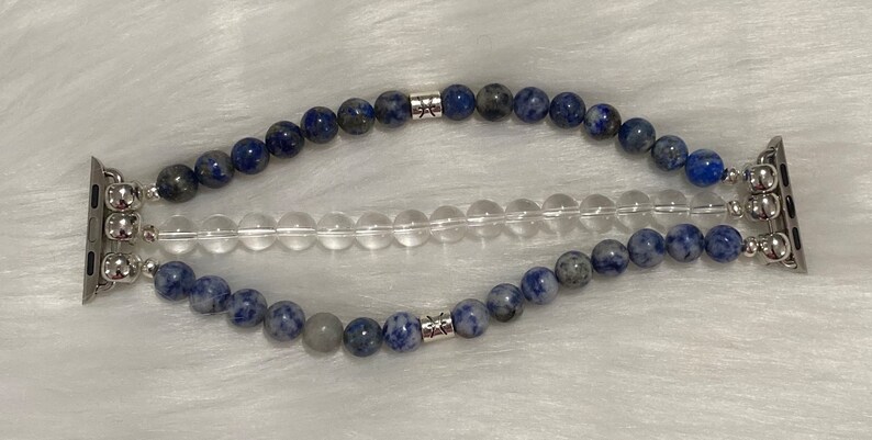 May include: A blue and gray beaded Apple Watch band with silver accents. The band has two rows of beads and a clear crystal row in the middle.