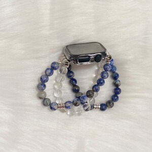 May include: A silver Apple Watch with a clear case is surrounded by three beaded bracelets. Two of the bracelets are made of blue and white stones, and the third is made of clear stones.