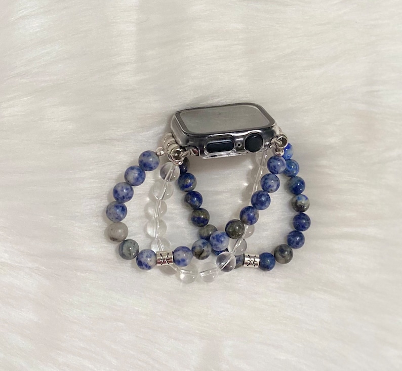 May include: A silver Apple Watch with a clear case is surrounded by three beaded bracelets. Two of the bracelets are made of blue and white stones, and the third is made of clear stones.