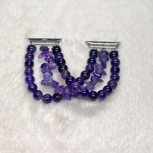 May include: A purple amethyst gemstone bracelet with a silver metal clasp. The bracelet is made of small, round beads and larger, irregular shaped beads. The bracelet is designed to fit an Apple Watch.