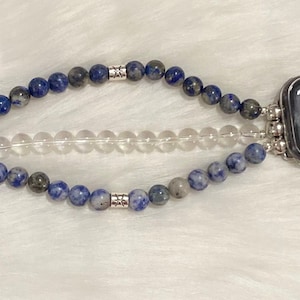 May include: A blue and gray beaded Apple Watch band with a clear crystal band layered on top. The band is attached to a silver Apple Watch.