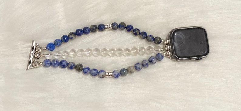 May include: A blue and gray beaded Apple Watch band with a clear crystal band layered on top. The band is attached to a silver Apple Watch.
