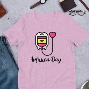 Może przedstawiać: Różowy T-shirt z grafiką przedstawiającą kroplówkę z sercem na końcu rurki. Pod grafiką znajduje się napis "Infusion Day".