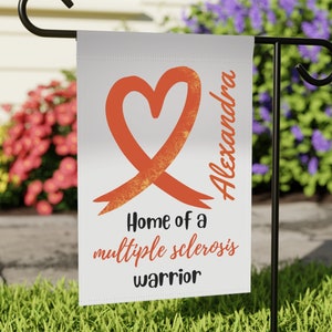 May include: A white garden flag with an orange heart and ribbon, the text "Home of a multiple sclerosis warrior" and the name "Alexandra".