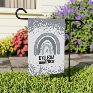 May include: A gray garden flag with a rainbow design and the text "Dyslexia Awareness" and a gray ribbon. The background has a white and gray splatter pattern.