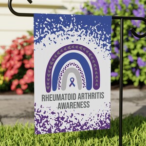 May include: A white garden flag with a purple, blue, and gray rainbow design. The rainbow has a purple ribbon in the center. The text "RHEUMATOID ARTHRITIS AWARENESS" is printed below the rainbow.