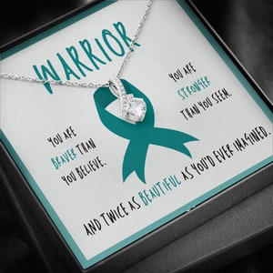 May include: A silver necklace with a pendant featuring a large, clear gemstone. The pendant is displayed on a card with a teal ribbon and the word "WARRIOR". Text on the card reads "You are braver than you believe" and "You are stronger than you seem."