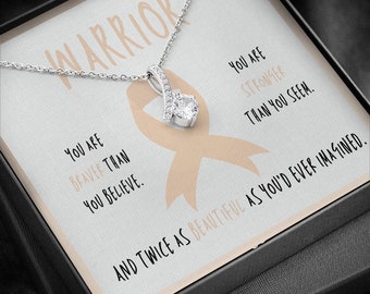 May include: A silver necklace with a pendant in the shape of a ribbon with a clear crystal in the center. The necklace is in a box with a card that reads "Warrior" and "You are braver than you believe. You are stronger than you seem. And twice as beautiful as you'd ever imagined."