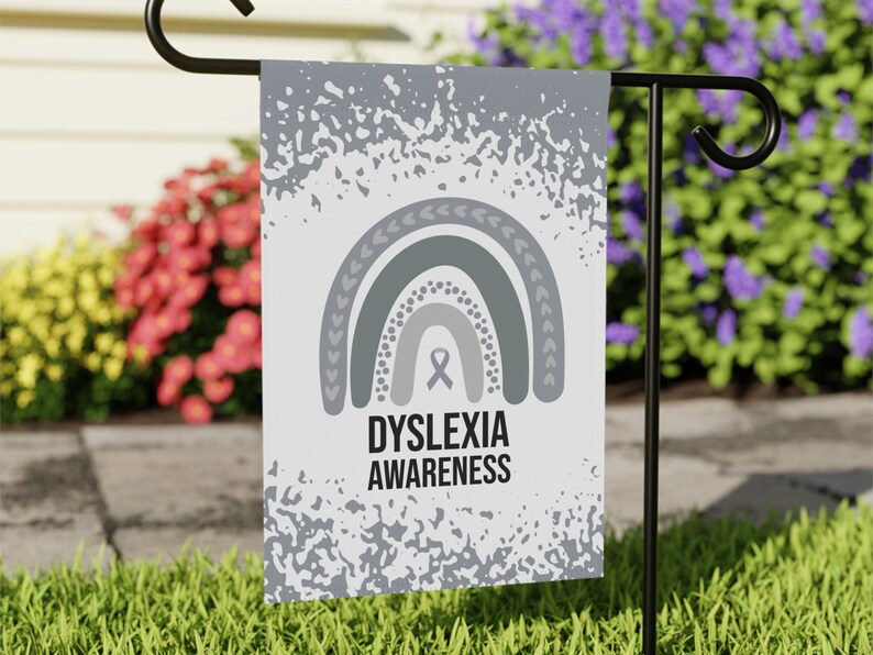 May include: A white garden flag with a gray and white rainbow design and the text "Dyslexia Awareness". The flag is hanging on a black metal flagpole.