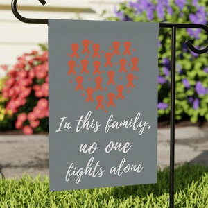 May include: A gray garden flag with a heart shape made of orange ribbons. The text "In this family, no one fights alone" is written below the heart.
