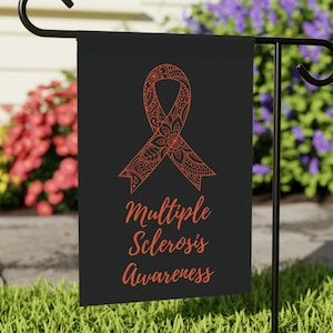 May include: A black garden flag with an orange awareness ribbon design and the words "Multiple Sclerosis Awareness" in a script font. The flag is displayed outdoors on a black metal stand, with a background of green grass and colorful flowers.