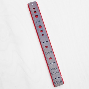 Könnte beinhalten: Ein hölzernes Spielbereich-Lineal mit roter Mitte und graviertem Text. Das Lineal ist segmentiert und mit "FAR", "CLOSE", "VERY CLOSE" und "MELEE" beschriftet. Es hat kleine kreisförmige Löcher und weiße Punkte zur Ausrichtung.