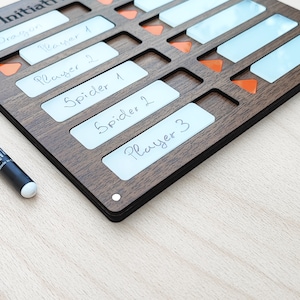 May include: A wooden initiative tracker with slots for player names and initiative order. The tracker has a dark brown finish, with white labels and orange markers. A black marker is visible on the left side. The text on the labels includes "Dragon", "Player 1", "Player 2", "Spider 1", "Spider 2", and "Player 3".