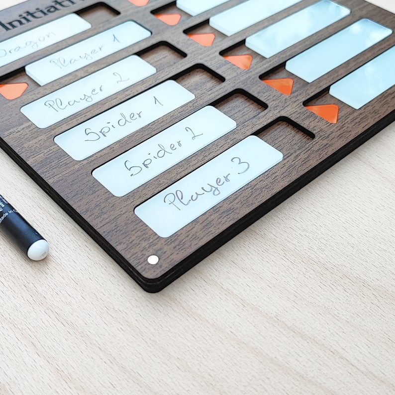 May include: A wooden initiative tracker with slots for player names and initiative order. The tracker has a dark brown finish, with white labels and orange markers. A black marker is visible on the left side. The text on the labels includes "Dragon", "Player 1", "Player 2", "Spider 1", "Spider 2", and "Player 3".