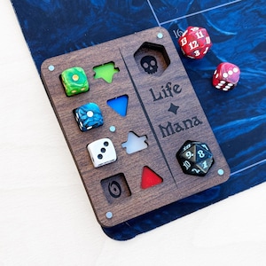 Peut inclure: Un plateau de jeu en bois avec des découpes pour dés et pions. Le plateau est gravé avec "Life Mana" et une icône de crâne. Plusieurs dés multicolores sont dispersés autour du plateau, dont rouges, verts, bleus et blancs.