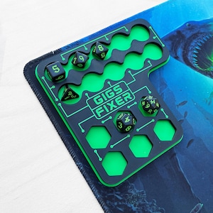 May include: A black and neon green dice tray featuring a circuit board pattern and the text GIGS FIXER. The tray has multiple circular and hexagonal cutouts for storing polyhedral dice, with five black dice with green numbers placed inside.