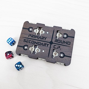 May include: A rectangular dark wood game tracker featuring four rotating dials with numbers 1, 8, 2, 0, 2, and 9 visible. The board is engraved with the text PRIMARY, SECONDARY, ROUND, and C.P. Three six-sided dice in blue, red, and blue-black are nearby.