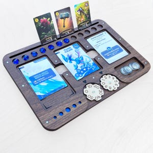 May include: A dark brown wooden game organiser with spaces for cards, dice, and tokens. It has blue accents. Several game cards are displayed, along with blue dice and wooden dials. The organizer is approximately 30 cm wide.
