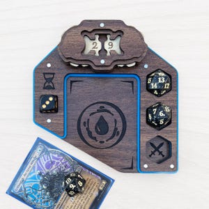 Könnte beinhalten: Ein hölzernes Spielzubehör mit dunkelbraunem Finish und blauem Rand. Es verfügt über Würfelhalter, einen Kartensteckplatz und einen Zähler mit den Zahlen 2 und 9. Eine Spielkarte und Würfel sind enthalten.