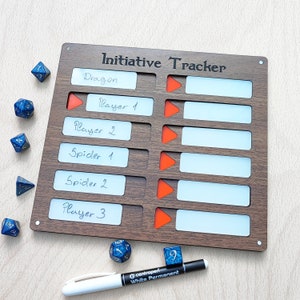 May include: A wooden initiative tracker with the text "Initiative Tracker" at the top. It has slots for names and tracking progress, with orange arrow indicators. Includes blue and white dice and a white permanent marker.