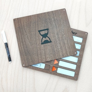 May include: A wooden board game with a dark brown finish, featuring an hourglass symbol and a section with white rectangles and orange triangles. A white marker pen is placed on the left side of the board.
