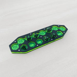 Puede incluir: Una bandeja de dados negra y verde con un diseño futurista. La bandeja tiene varias ranuras hexagonales y recortes circulares, con los números "DG" y "D8" visibles. Varios dados están colocados en la bandeja. La bandeja está hecha de acrílico.