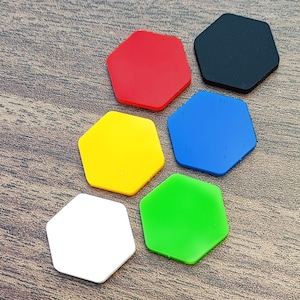 May include: Six colorful, hexagonal shapes in red, black, blue, green, yellow, and white are arranged on a wooden surface. The shapes are solid and evenly colored, with a smooth, matte finish. The arrangement suggests a game or craft component.