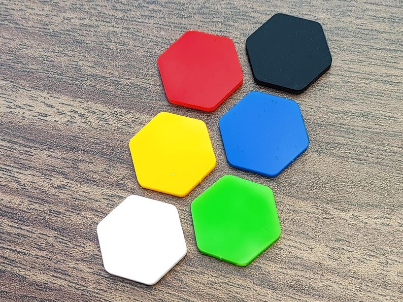 May include: Six colorful, hexagonal shapes in red, black, blue, green, yellow, and white are arranged on a wooden surface. The shapes are solid and evenly colored, with a smooth, matte finish. The arrangement suggests a game or craft component.