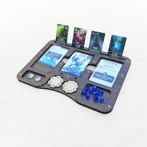 Könnte beinhalten: Ein Spiel-Organizer aus dunklem Holz mit Schlitzen für Karten, Würfel und Spielsteine. Der Organizer enthält mehrere Spielkarten mit Illustrationen und Text, sowie blaue Würfel, weiße Zifferblätter und kleine runde Spielsteine. Die Karten zeigen Fantasy-Kunst.