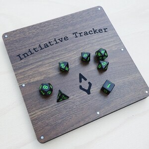 May include: A wooden Initiative Tracker board with the text "Initiative Tracker" in black. The board features a dark wood grain pattern and includes several dice with green numbers and markings. A black arrow symbol is also present.