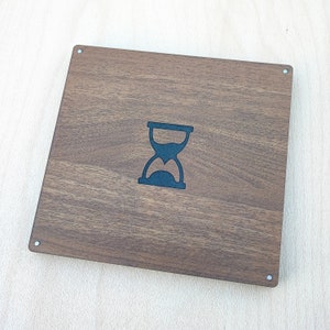 May include: A square, wooden box with a dark brown finish. The box features a black hourglass design in the center. The corners have small, silver-colored rivets. The wood grain is visible.