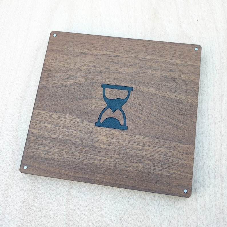 May include: A square, wooden box with a dark brown finish. The box features a black hourglass design in the center. The corners have small, silver-colored rivets. The wood grain is visible.