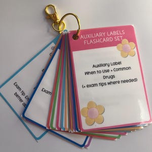 May include: A set of flashcards with a gold-colored ring and clasp. The top card is pink with the text "AUXILIARY LABELS FLASHCARD SET" and a flower graphic. The cards have colorful edges and include text about auxiliary labels and exam tips.