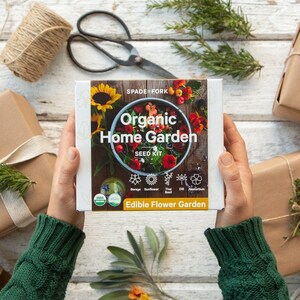 May include: A person holding a white box with the text "Organic Home Garden Seed Kit" and "Edible Flower Garden". The box is surrounded by brown paper wrapped gifts with twine and flowers.