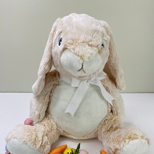 May include: A plush beige bunny rabbit with a white bow sits on a white surface surrounded by pastel colored speckled Easter eggs and orange carrot decorations.