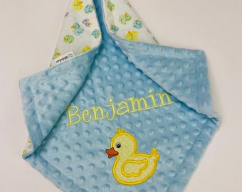 May include: A light blue baby blanket with a yellow rubber ducky and the name "Benjamin" embroidered on it. The blanket is made of soft, plush fabric and has a white side with a pattern of yellow ducks.