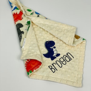 May include: A white and cream colored baby blanket with a dinosaur print on one side and a white side with a blue dinosaur and the name "Brogan" embroidered on it.