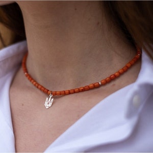 May include: A necklace featuring a strand of small, cylindrical orange beads with a silver-toned metal pendant shaped like the Ukrainian Tryzub emblem. The beads are uniform in size and color, interspersed with tiny silver-toned spacers along the strand.