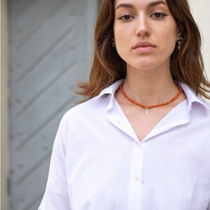 May include: A white button-down shirt with a collar and a beaded necklace featuring small orange spherical beads and a silver-toned pendant shaped like a trident symbol, worn around the neck against the fabric of the shirt.