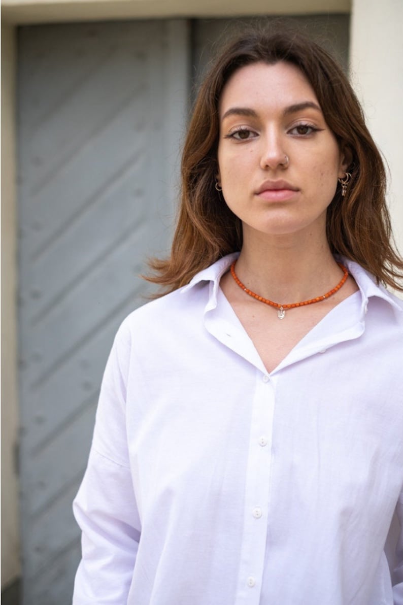 May include: A white button-down shirt with a collar and a beaded necklace featuring small orange spherical beads and a silver-toned pendant shaped like a trident symbol, worn around the neck against the fabric of the shirt.