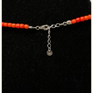 May include: A necklace featuring small, vibrant orange cylindrical beads strung together. The piece includes a silver-toned metal lobster claw clasp, an adjustable chain link extender, and a small round tag clearly stamped with the number 925.