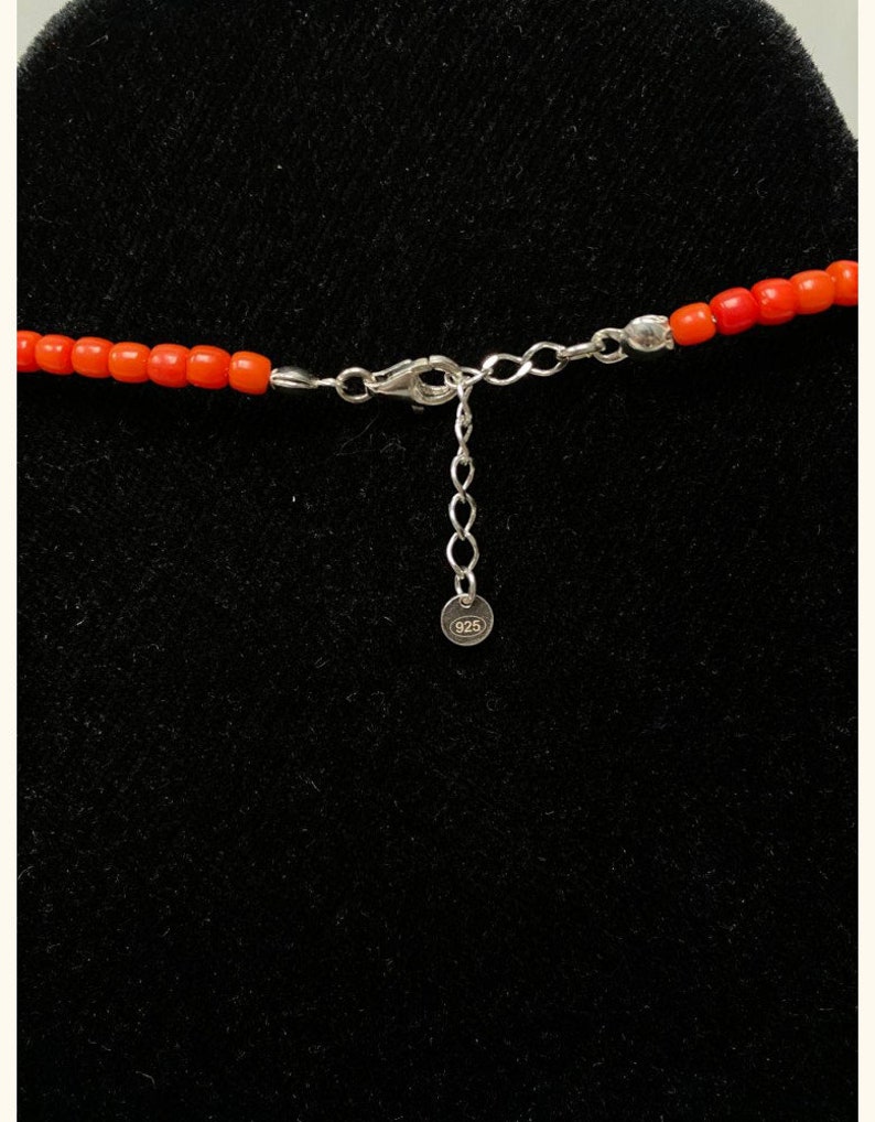 May include: A necklace featuring small, vibrant orange cylindrical beads strung together. The piece includes a silver-toned metal lobster claw clasp, an adjustable chain link extender, and a small round tag clearly stamped with the number 925.
