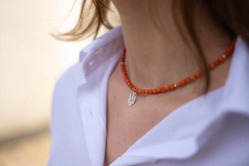 May include: A necklace featuring small, round, orange beads strung together with occasional silver-toned spacers. A silver-toned pendant shaped like the Ukrainian Tryzub emblem hangs from the center of the beaded strand.