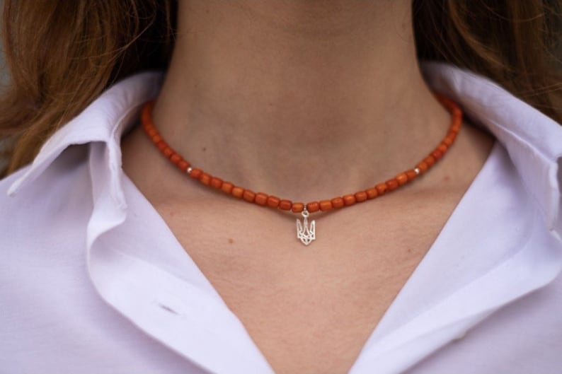 May include: A choker necklace featuring small, cylindrical orange beads strung together with a central silver-toned pendant shaped like the Ukrainian Tryzub emblem. The necklace includes small silver-toned spacer beads along the strand.