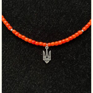 May include: A necklace featuring a string of small, vibrant orange beads interspersed with tiny silver-toned spacers. A silver-toned metal pendant in the shape of a trident symbol hangs from the center of the beaded strand.