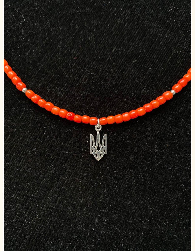 May include: A necklace featuring a string of small, vibrant orange beads interspersed with tiny silver-toned spacers. A silver-toned metal pendant in the shape of a trident symbol hangs from the center of the beaded strand.