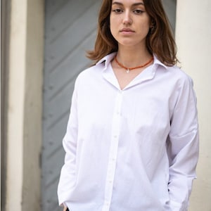 May include: A crisp white long-sleeved button-down shirt with a classic collar and a front button placket, paired with black trousers and an orange beaded necklace featuring a small silver pendant charm resting at the base of the neck.