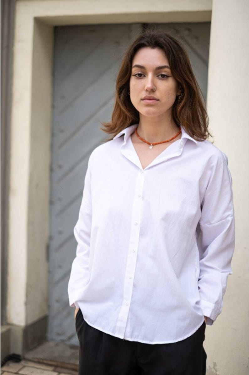 May include: A crisp white long-sleeved button-down shirt with a classic collar and a front button placket, paired with black trousers and an orange beaded necklace featuring a small silver pendant charm resting at the base of the neck.